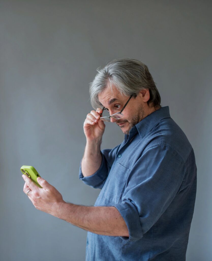 Mature man adjusting glasses while checking smartphone. Gray-haired man views green device against neutral wall.
